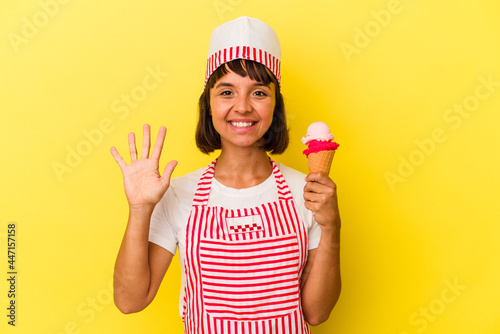 Young mixed race ice cream maker woman holding an ice cream isolated on yellow background smiling cheerful showing number five with fingers. © Asier