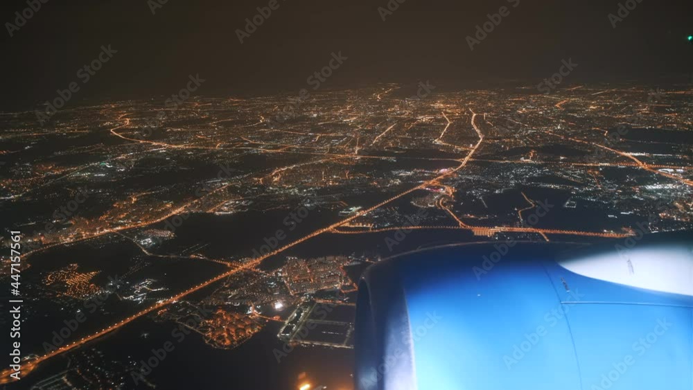 View from the plane window of the lights of the glowing city at night and the plane's engine on the wing. Flight over the metropolis at night on an airplane view from the wing window.