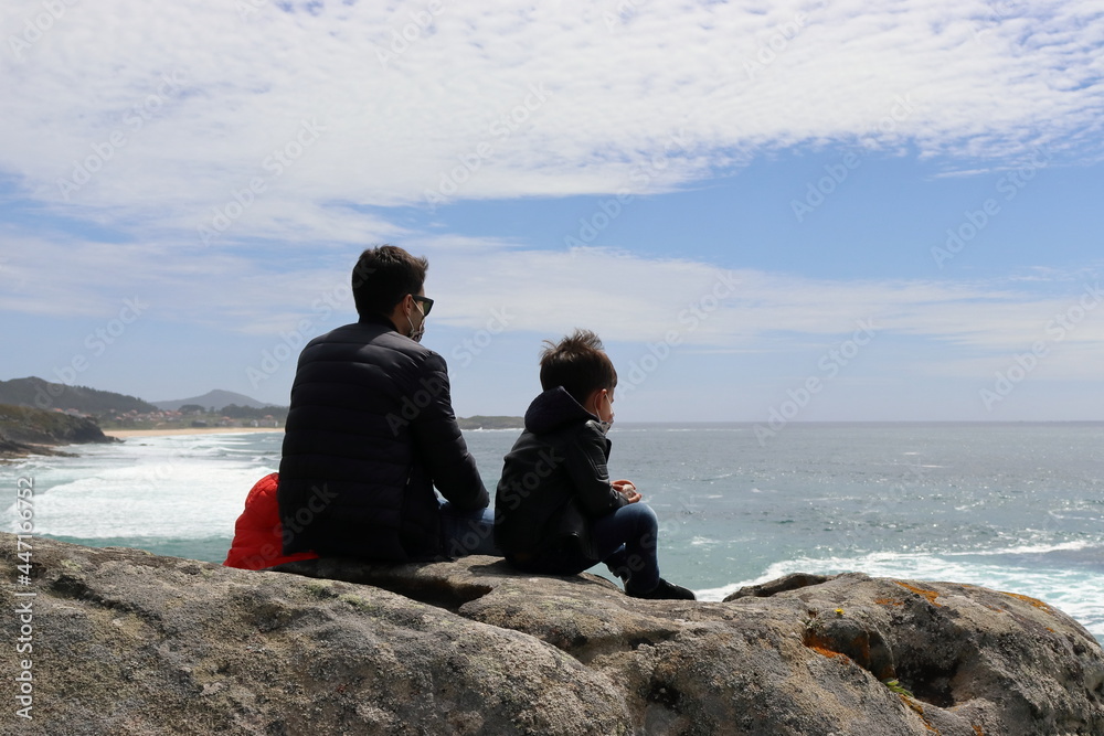Father and young son sitting on a rock looking out to sea.