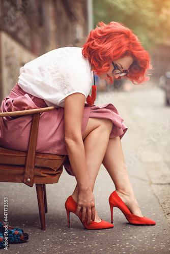 Fashionable young girl with red hair and red shoes sitting in an urban part of the city, checking her shoe