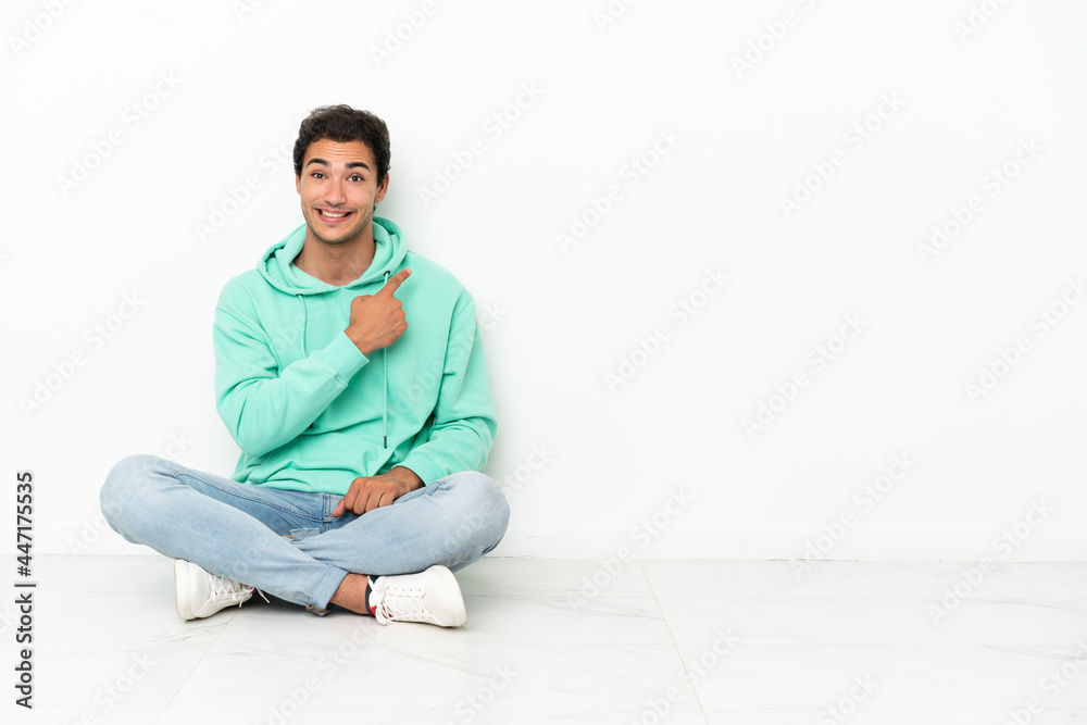 Caucasian handsome man sitting on the floor pointing to the side to present a product Caucasian handsome man sitting on the floor pointing to the side to present a product