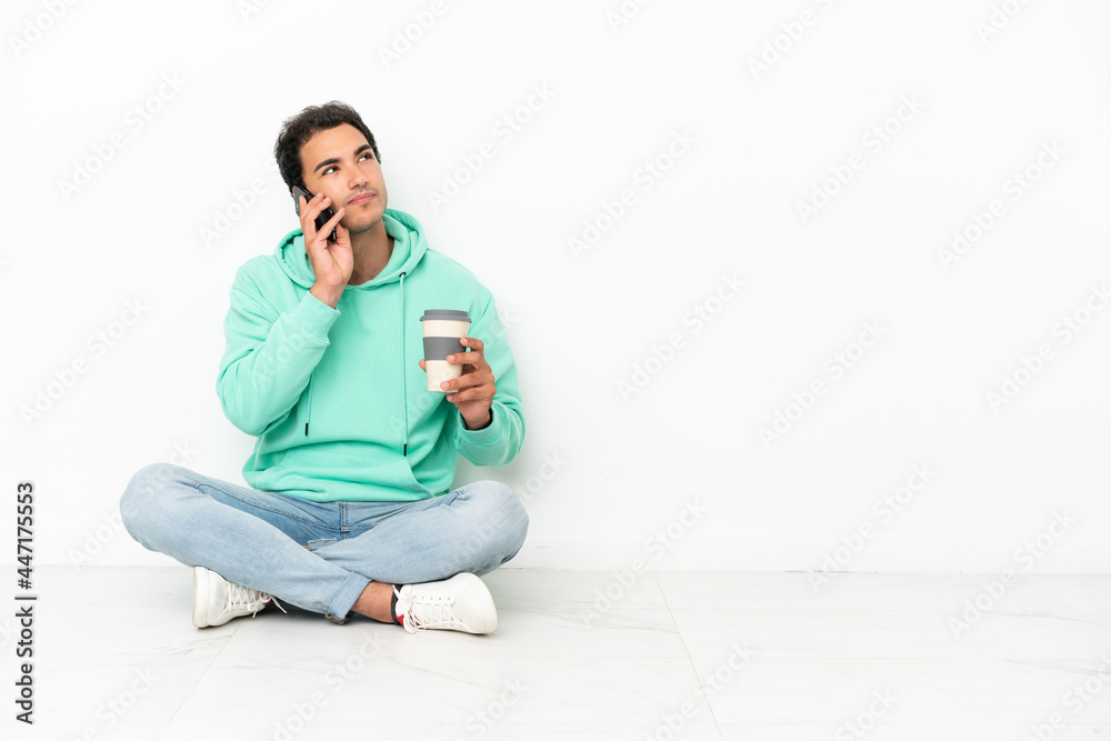 Caucasian handsome man sitting on the floor holding coffee to take away and a mobile