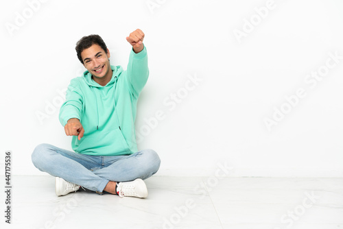 Caucasian handsome man sitting on the floor points finger at you while smiling