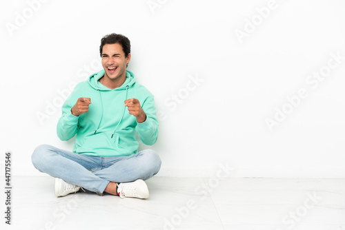 Caucasian handsome man sitting on the floor pointing to the front and smiling