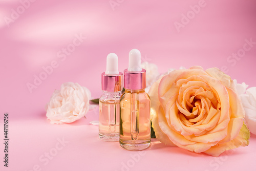 Essential rose oil in cosmetic bottle near fresh rose flowers against pink background.