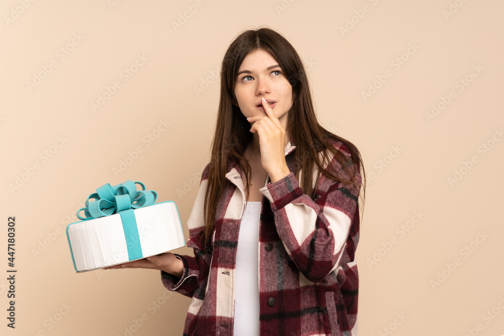 Young Ukrainian girl holding a big cake isolated on beige background having doubts while looking up Young Ukrainian girl holding a big cake isolated on beige background having doubts while looking up