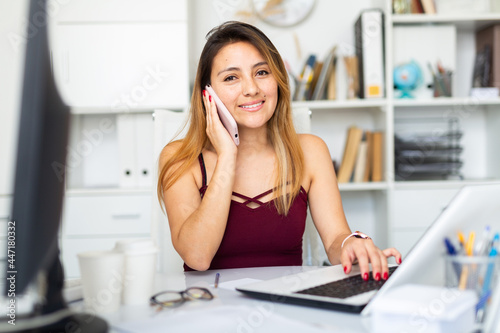 Positive latina woman in casual wear using mobile phone while working alone with laptop and papers in office © JackF