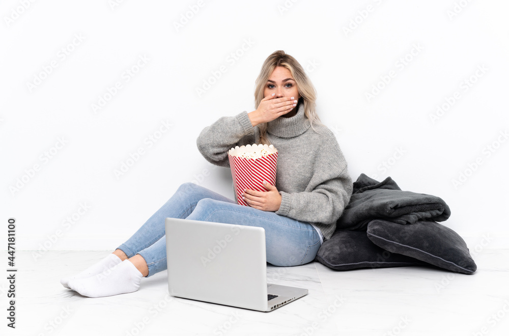 Teenager blonde girl eating popcorn while watching a movie on the laptop covering mouth with hands Teenager blonde girl eating popcorn while watching a movie on the laptop covering mouth with hands