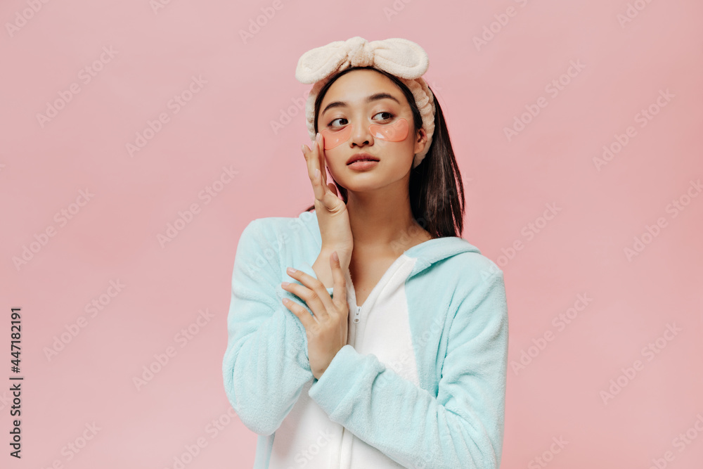 Brown-eyed brunette woman in blue pajamas and soft headband gently touches face and poses with cosmetic eyepatches on pink background. Brown-eyed brunette woman in blue pajamas and soft headband gently touches face and poses with cosmetic eyepatches on pink background.