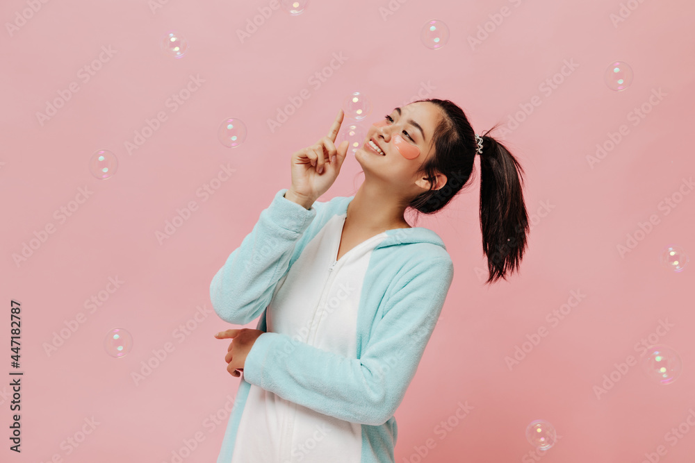 Pretty cool girl in blue kigurumi smiles, plays with bubbles and poses with cosmetic eyepatches on pink isolated background.