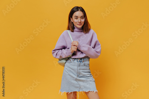 Young cool girl in denim skirt, stylish purple sweater and eyeglasses looks into camera, poses on orange background and holds mint backpack.