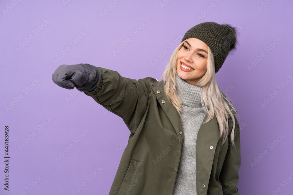 Teenager blonde girl with winter hat over isolated purple background giving a thumbs up gesture