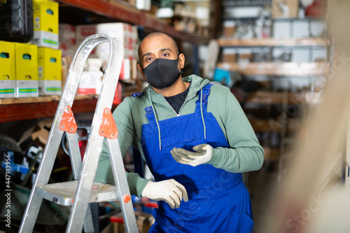 Warehouse worker in protective mask stands next to stepladder and tool shelves in a hardware store