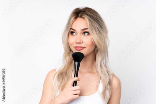 Teenager blonde girl over isolated white background holding makeup brush and thinking