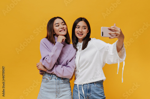 Cool young brunette Asian women in stylish sweatshirts take selfie, smile sincerely and pose in good mood on orange background.