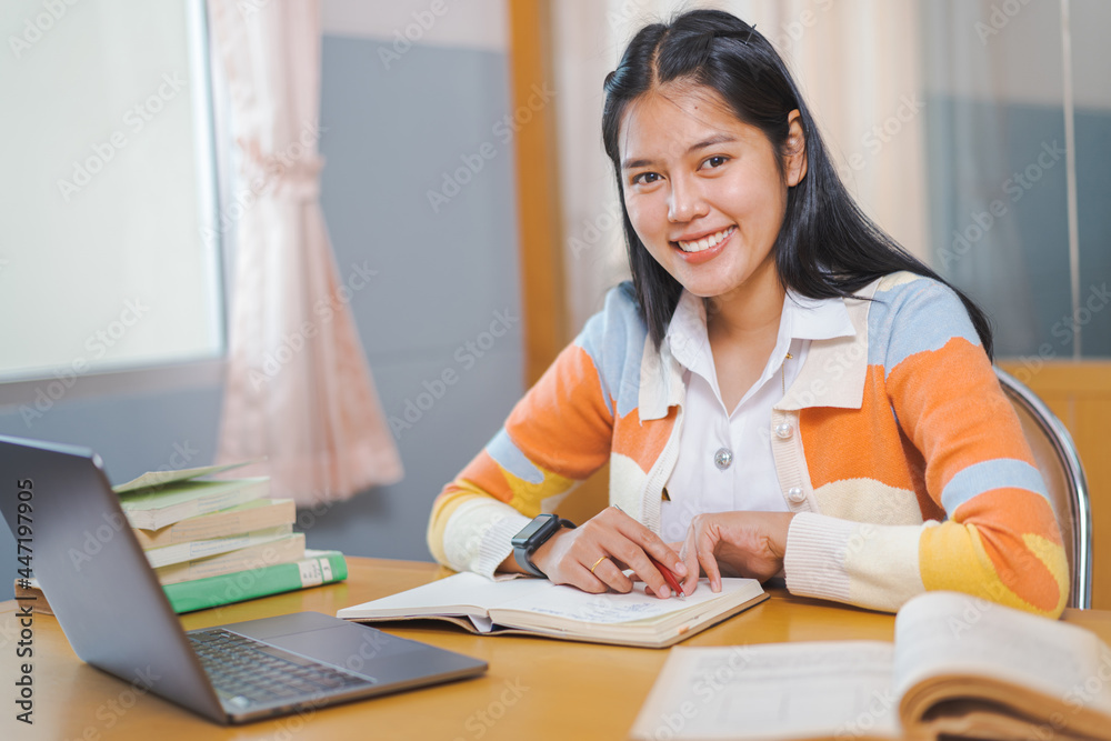 A young happy woman Asian college student in student uniform studying online, reading a book, using digital tablet or laptop in university library in free time by oneself A young happy woman Asian college student in student uniform studying online, reading a book, using digital tablet or laptop in university library in free time by oneself