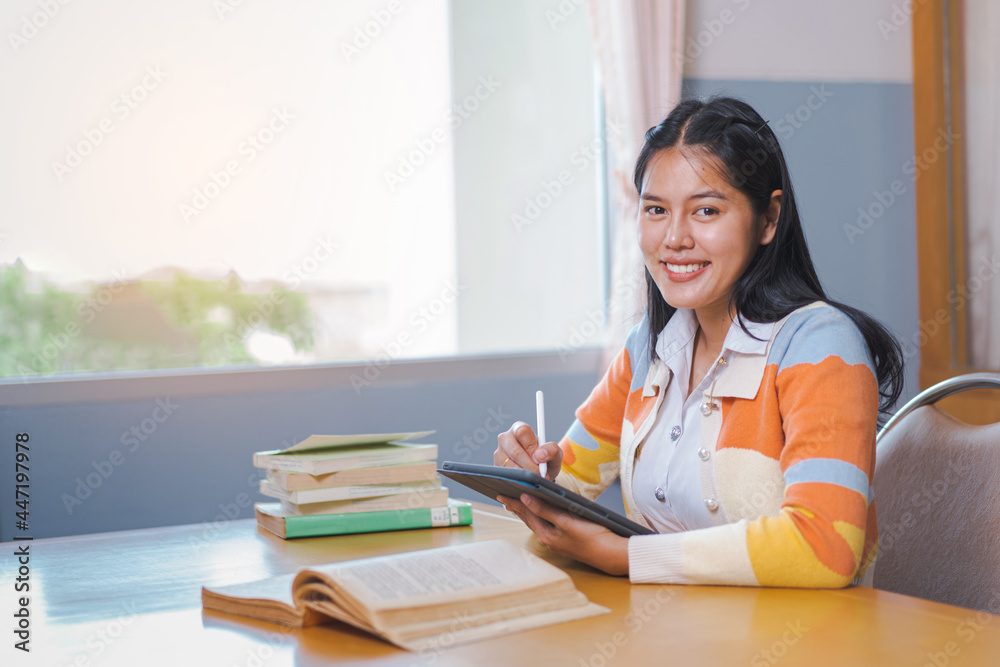A young happy woman Asian college student in student uniform studying online, reading a book, using digital tablet or laptop in university library in free time by oneself A young happy woman Asian college student in student uniform studying online, reading a book, using digital tablet or laptop in university library in free time by oneself