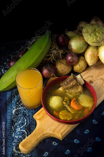 overhead view of a traditional Latin American dish of beef soup with vegetables and spices next to a glass of natural passion fruit soft drink on a traditional Costa Rican tablecloth