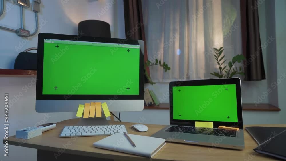 Laptop Computer and Desktop Computer Showing Green Key Screen Stands on a Desk in the Living Room Smooth Zoom in. In the Background Cozy Living Room in the Evening with Warm Lights on. Laptop Computer and Desktop Computer Showing Green Key Screen Stands on a Desk in the Living Room Smooth Zoom in. In the Background Cozy Living Room in the Evening with Warm Lights on.