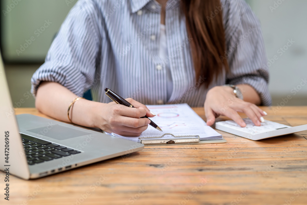 Image of a businesswoman holding a pen and using a calculator documents and laptop are placed on the table.