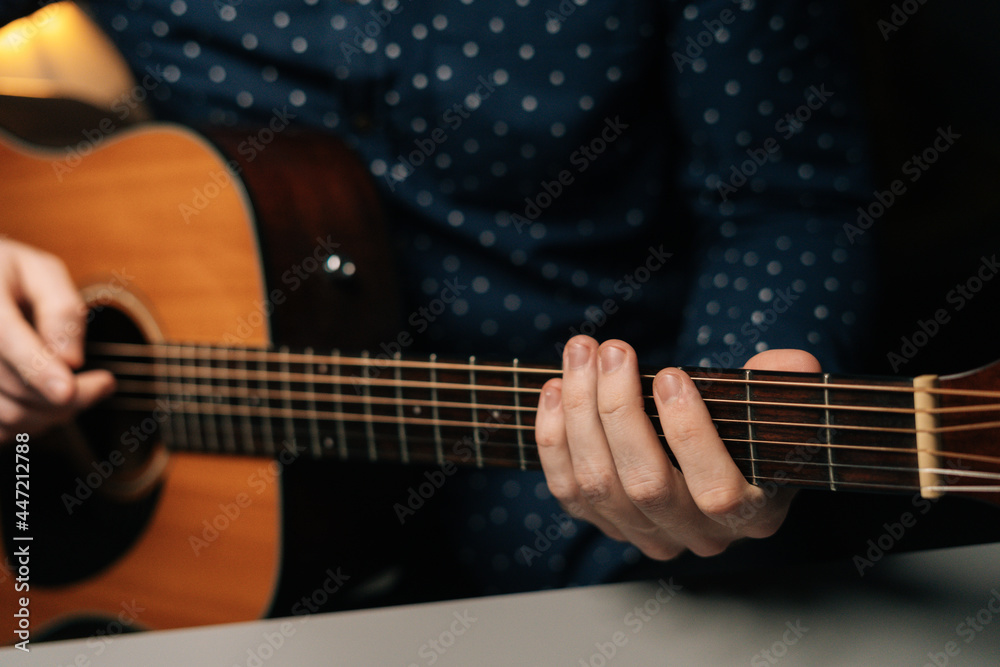 Close-up front view of unrecognizable guitarist male playing acoustic guitar sitting at desk in dark living room, selective focus. Creative musician enjoying leisure activity in apartment.