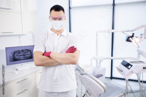 Portrait of a young dentist in protective mask, work glasses and in a white uniform stands in the office during coronavirus quarantine. Emergency care