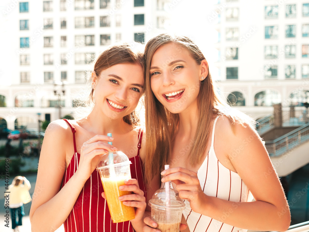 Two young beautiful smiling hipster female in trendy summer clothes. Carefree women posing outdoors.Positive models holding and drinking fresh cocktail smoothie drink in plastic cup with straw Two young beautiful smiling hipster female in trendy summer clothes. Carefree women posing outdoors.Positive models holding and drinking fresh cocktail smoothie drink in plastic cup with straw