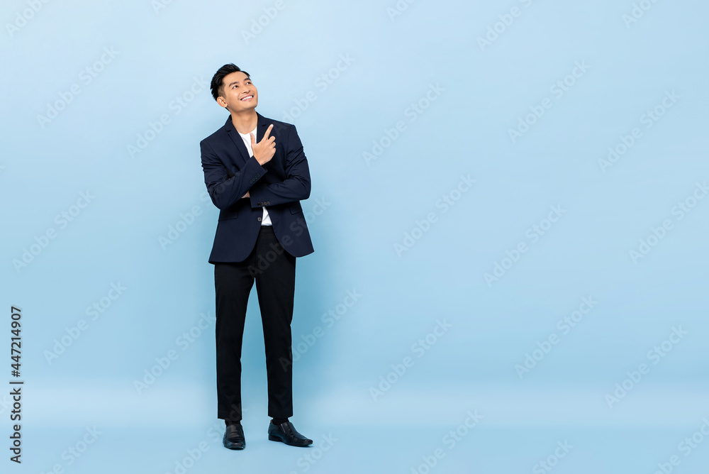 Full length portrait of young handsome southeast Asian businessman looking up and pointing to copy space on light blue studio background Full length portrait of young handsome southeast Asian businessman looking up and pointing to copy space on light blue studio background