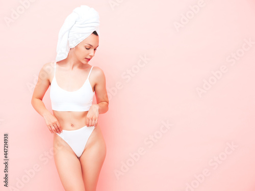 Young beautiful smiling woman in white lingerie. Sexy carefree model in underwear and towel on head posing pink wall in studio. Positive and happy female enjoying morning