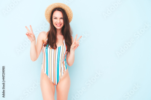 Young beautiful sexy smiling  hipster woman in posing near light blue wall in studio. Trendy model in colorful summer swimwear bathing suit. Positive model going crazy in hat.Shows peace sign