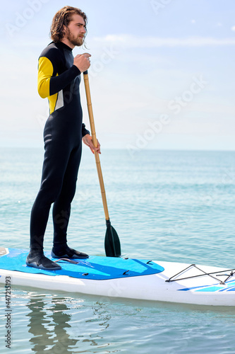 Bearded man with long hair in black wetsuit working out with stand up paddle board on water or in open sea,athelte caucasian guy is engaged in water sport, extreme sport. subsurfing concept
