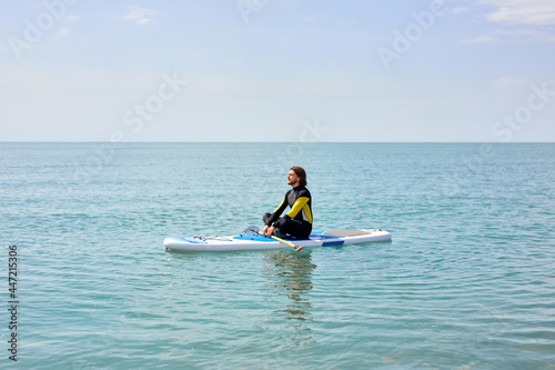 Side view portrait of calm bearded guy sitting on paddleboard meditating after sup surfing, in black wetsuit, keep calm, yoga and sport concept. copy space © Roman
