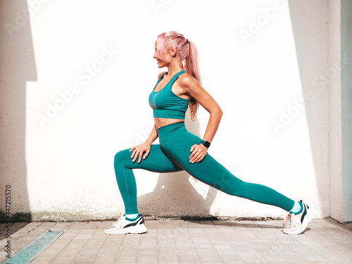 Fitness smiling woman in green sports clothing with pink hair. Young beautiful model with perfect body.Female posing in the street near white wall.Cheerful and happy. Stretching out before training
