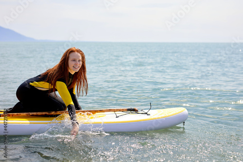 Young beuatiful redhead surfer woman making preparation for surf session, young lady in black swimsuit sail from the shore in sea, side view portrait. copy space