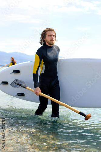 Photography young male in diving suit spedting time at sea, carrying surfboard and paddle in hands, after surfing alone, wearing black wetsuit