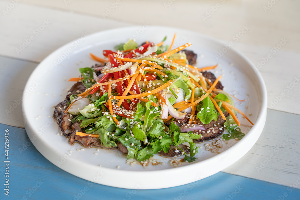 Fresh summer salad made of roasted beef meat slices, lettuce, grated carrot, sweet pepper and onion decorated with sesame seeds served on white round plate on striped wooden background at restaurant