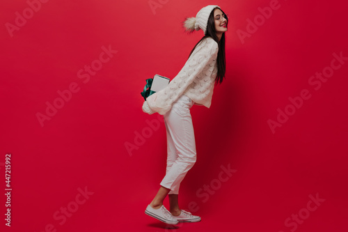 Full-length photo of trendy young brunette in white jeans, sneakers, knitted jersey and hat, smiling with closed eyes, preparing gift box isolated on red plain background 