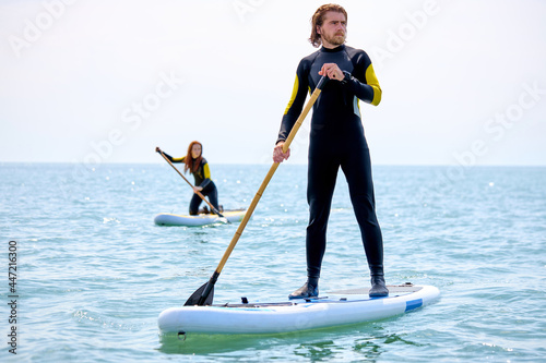 Fit sportive man and woman in wetsuit doing stand-up paddleboard in ocean, engaged in sport. Athlete paddleboarding on SUP surf board. Copy space. Focus on bearded guy with paddles