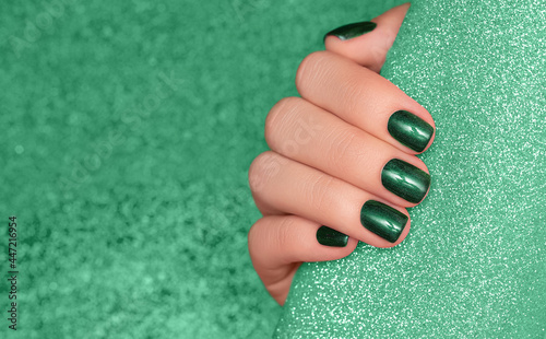 Female hand with green nail design. Glitter green nail polish manicure. Woman hand on glitter green fabric background