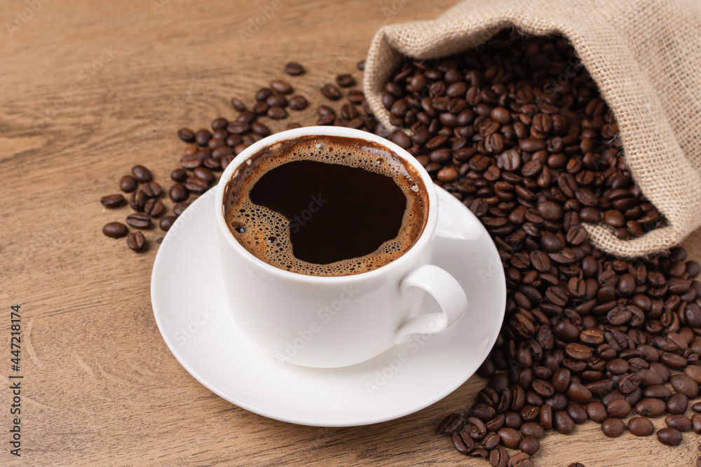 Fototapeta premium Coffee cup and saucer on wooden background with roasted coffee beans in a burlap sack.