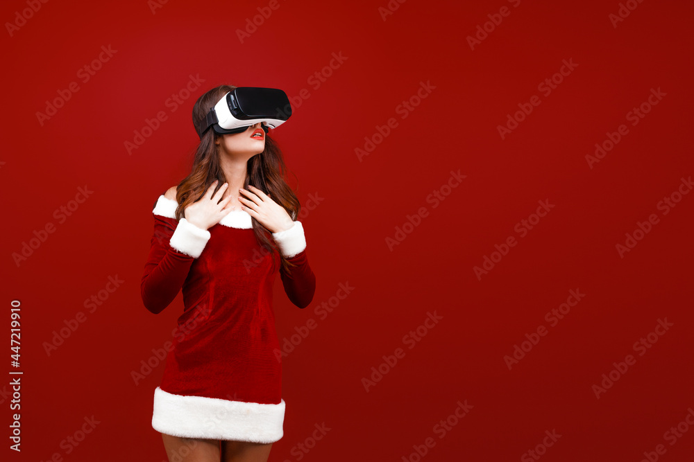 Obraz premium Amazed young Santa girl in Christmas dress looking in headset pointing hands aside isolated on red wall background.