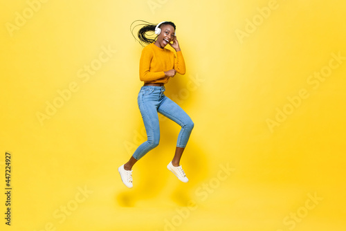 Happy energetic young African American woman wearing headphones listening to music and jumping in yellow isolated studio background
