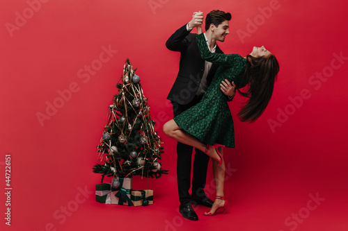 Fotografie Stylish brunette-haired gentleman in black suit smiling and dancing with lady in green dress and trendy high heels shoes