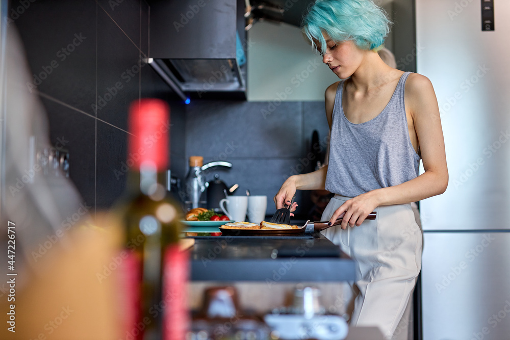 Attractive young woman with modern green hairstyle is cooking breakfast, side view. Beautiful caucasian lady in casual domestic clothes preparing meal in the morning, copy space. side view portrait