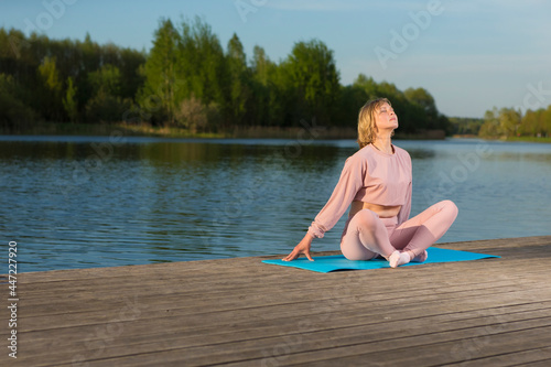 Female Sport Concepts. Relaxing Senior Woman in Sport Outfit Practicing Yoga On Wooden Stage Near Water With Closed Eyes Outdoor. © danmorgan12