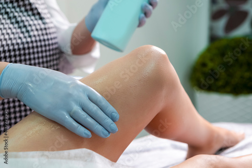 Bodycare Ideas. Beautician During Wax Depilation Procedure of Legs By Wax Device on Female Legs in Beauty Salon.