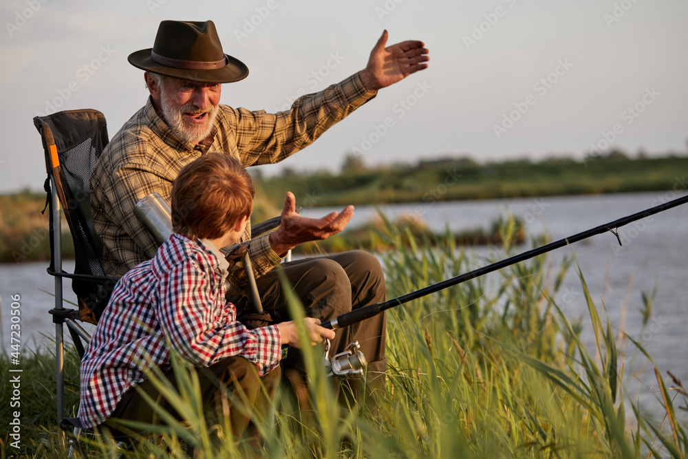 Grandfather and kid boy together fishing in the evening at sunset time in summer day on river in countryside, side view on caucasian multi-generation family, elderly man teach child boy to fish - obrazy, fototapety, plakaty 