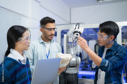 Caucasian male engineer standing hold a robot controller next to Asian female apprentice holding a laptop, demonstrating the work of robotic machine to young Asian male CEO who smile touching a robot.