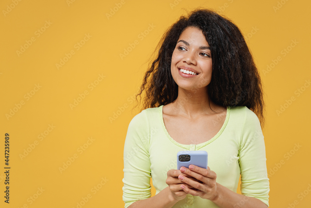 Joyful fun charming fascinating overjoyed african american young brunette woman 20s wears green shirt hold in hand use mobile cell phone look aside away isolated on yellow background studio portrait. Joyful fun charming fascinating overjoyed african american young brunette woman 20s wears green shirt hold in hand use mobile cell phone look aside away isolated on yellow background studio portrait.
