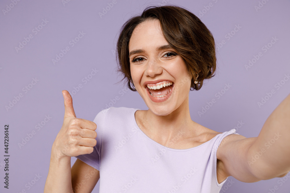 Close up young smiling happy friendly woman 20s with bob haircut wear white t-shirt doing selfie shot on mobile phone show thumb up like gesture isolated on pastel purple background studio portrait. Close up young smiling happy friendly woman 20s with bob haircut wear white t-shirt doing selfie shot on mobile phone show thumb up like gesture isolated on pastel purple background studio portrait.